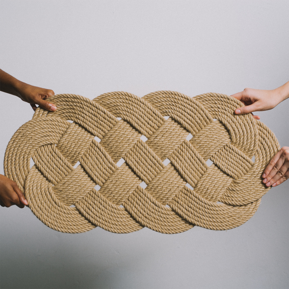 Three hands holding a thickly braided tan rope mat against a plain white background.
