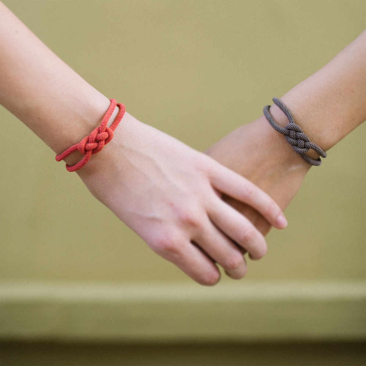 Two hands wearing knotted rope bracelets in red and gray against a neutral background.