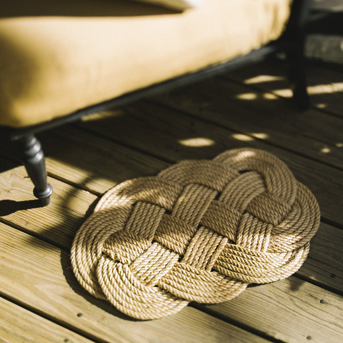 Woven rope doormat on wooden deck beside cushioned bench, natural outdoor decor