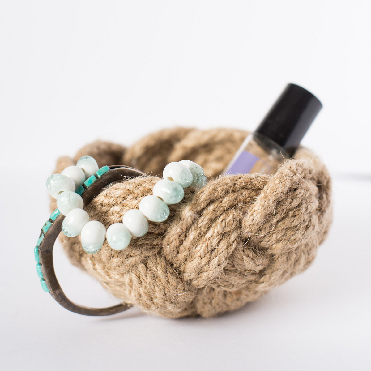 Braided rope basket holding beaded bracelet and small bottle on white background